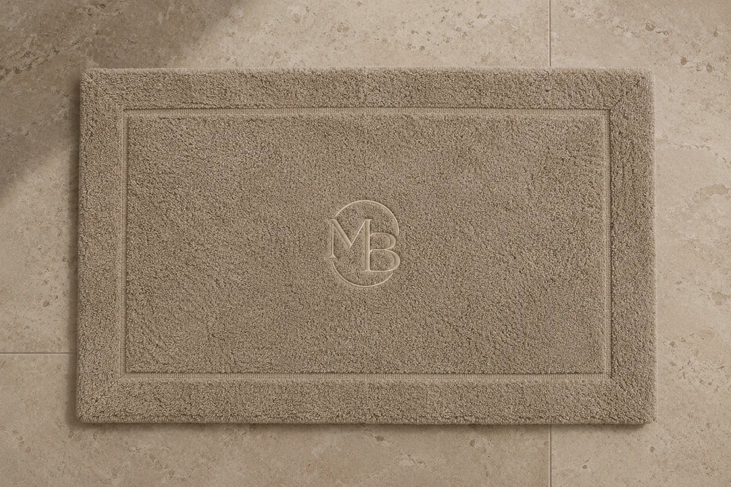 Tapis de bain MDB taupe posé sur un sol en pierre, monogramme au centre
